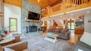 A spacious living room with a stone fireplace, a large TV mounted above it, and a wooden balcony. - Dog Friendly Lakefront Home w/ Pool & Hot Tub! (Swanton)