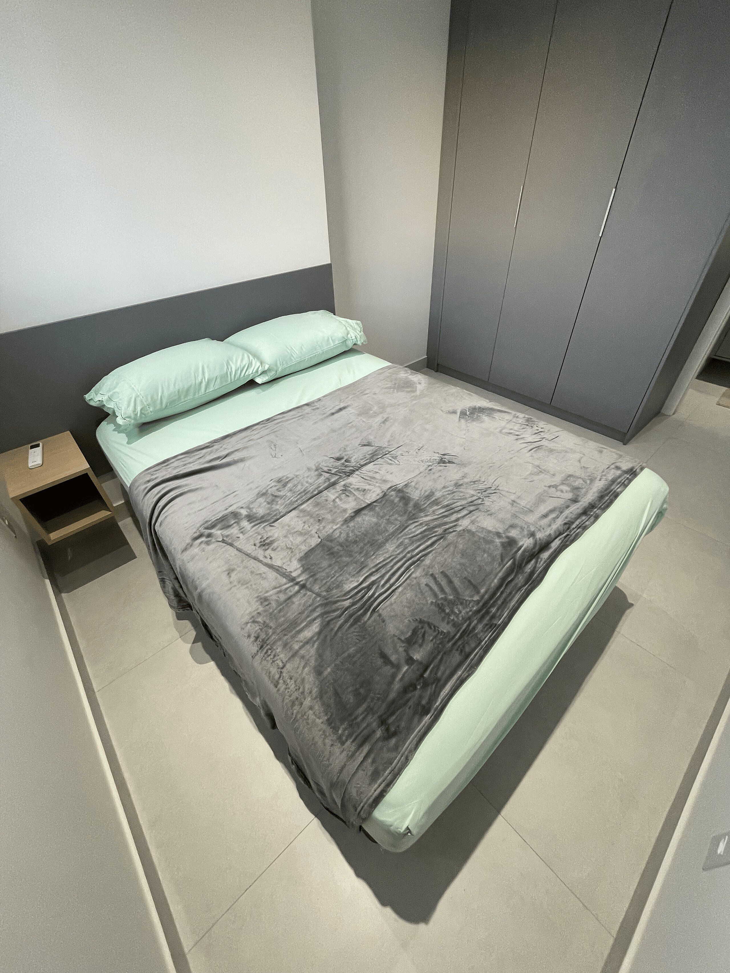 Sängyssä on harmaa ja vihreä päiväpeite, vaaleanvihreä tyyny ja puinen yöpöytä, jonka päällä on puhelin.