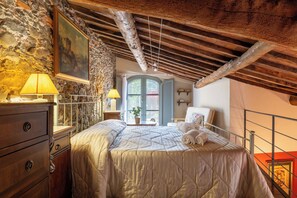 A bedroom with a stone wall, a bed with a quilt, a wooden dresser, a nightstand, a lamp, and a painting. - Superior Apartment Borgo di Matraia (Matraia)
