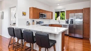 A modern kitchen with a large island, wooden cabinets, stainless steel appliances, and black bar stools. - Newly Remodeled Home in Waikoloa Village!  WeloWelo House (Waikoloa Village)