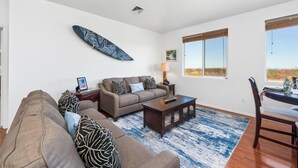 A living room with a sofa, coffee table, and a dining table with chairs. There is a surfboard on the wall and a window with a view. - Newly Remodeled Home in Waikoloa Village!  WeloWelo House (Waikoloa Village)