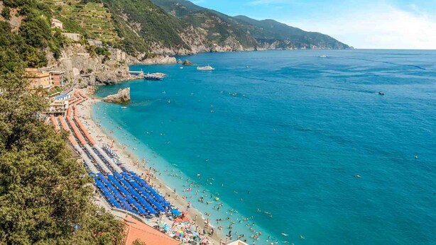 Ein Küstenstrand mit freiem Blick auf das Meer, einer felsigen Uferlinie und einem Hügel mit Gebäuden.