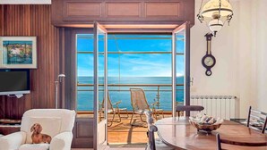 Ein Zimmer mit einer großen Schiebeglasdoor zum Balkon mit Meerblick. Es gibt einen Esstisch mit Stühlen, einen Fernseher und eine Wanduhr.