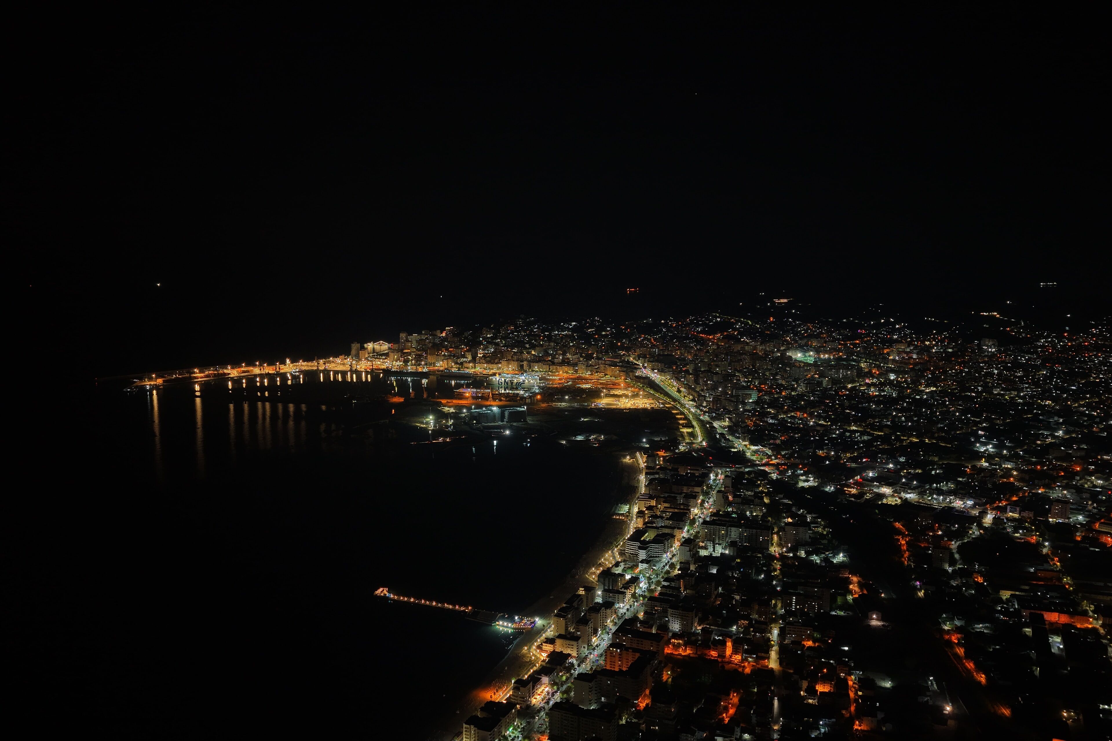 An aerial view of a coastal city at night with illuminated streets and a prominent bridge.