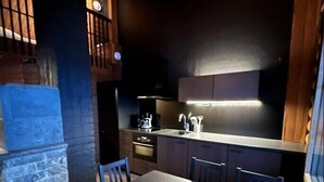 Cozinha moderna com bancada escura, armários de madeira e forno embutido. Há uma mesa de jantar com cadeiras e uma escada que leva a um andar superior.