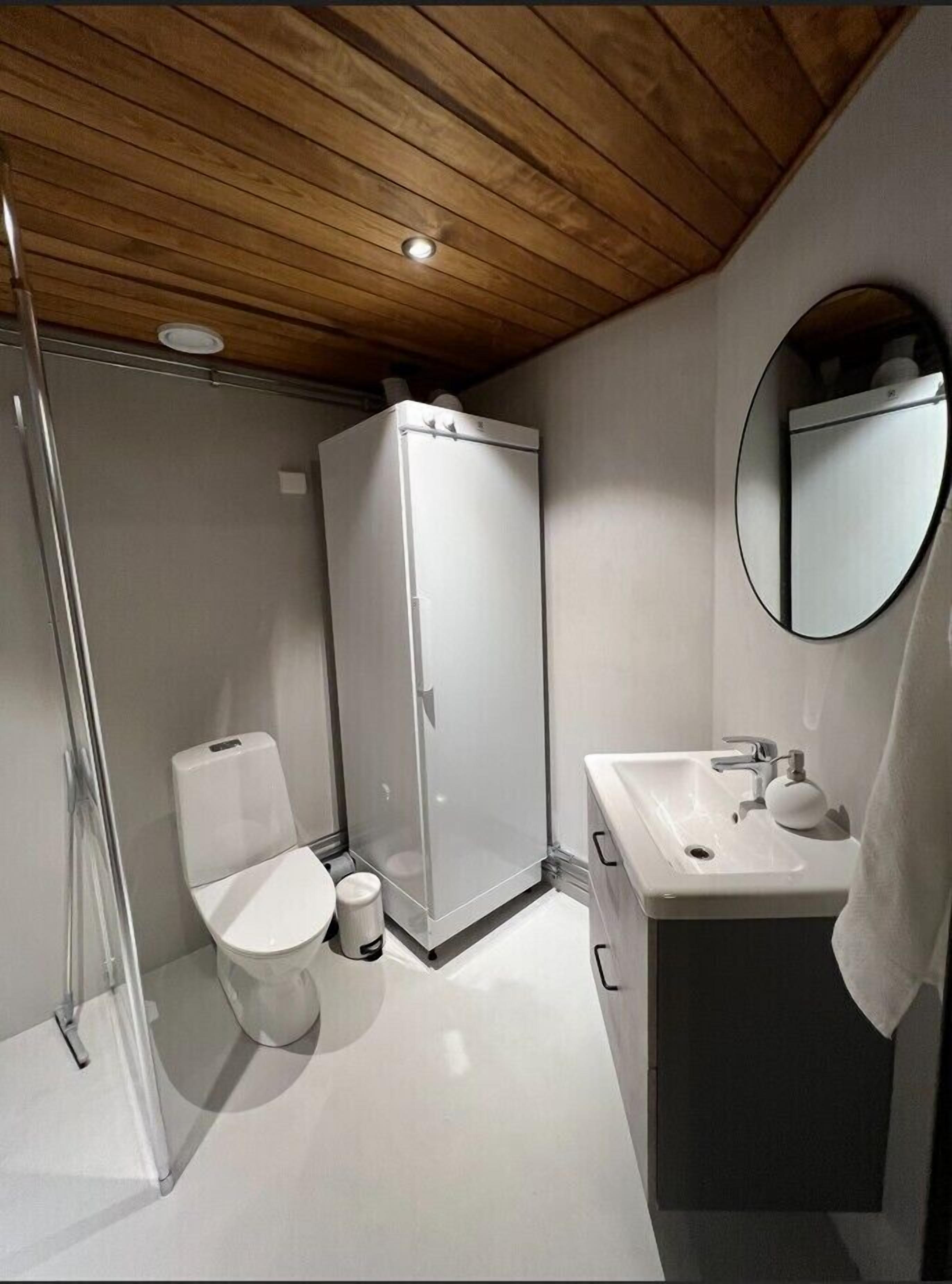 Une salle de bain moderne avec un plafond en bois, une cabine de douche en verre, des toilettes, un lavabo avec un miroir et une poubelle.