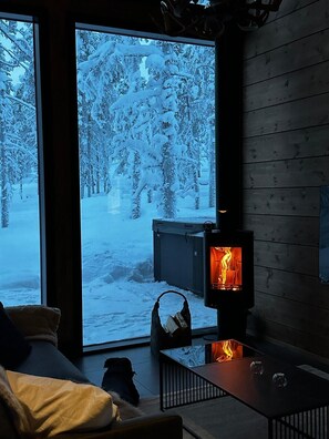 Une pièce chaleureuse avec une grande fenêtre donnant sur une forêt enneigée, une cheminée et un coin salon confortable.