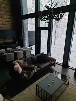 A modern living room with a large window, a leather sofa, a glass coffee table, and a chandelier.