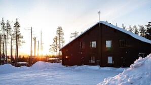 A wooden cabin with a sloped roof, surrounded by snow and trees.
