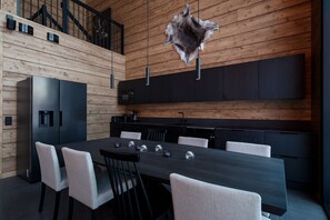 A modern kitchen with wooden panel walls, a black countertop, and a dining area with a long table and chairs. - Levi utsuvaara frame b (Sirkka)