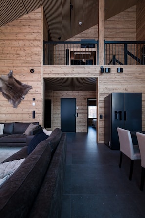 A spacious interior with a high ceiling, wooden walls, a large sofa, a dining area, and a staircase leading to an upper level.