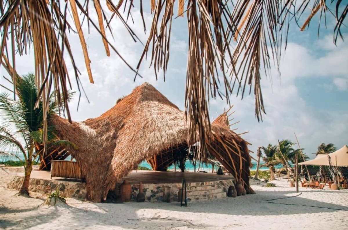 A thatched-roof structure on a sandy beach with palm trees and a clear sky.
