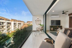 A modern balcony with a glass railing, a seating area with a sofa and chairs, a flat-screen TV, and a plant. - Ts'íibol: 3BR w Private Rooftop Deck & Pool (Tulum)