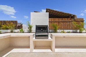 A modern outdoor seating area with a grill, surrounded by a concrete structure and bamboo fencing, with a thatched-roof building in the background. - Sleeps 6 w Private Rooftop Pool & 2 Bikes (Tulum)