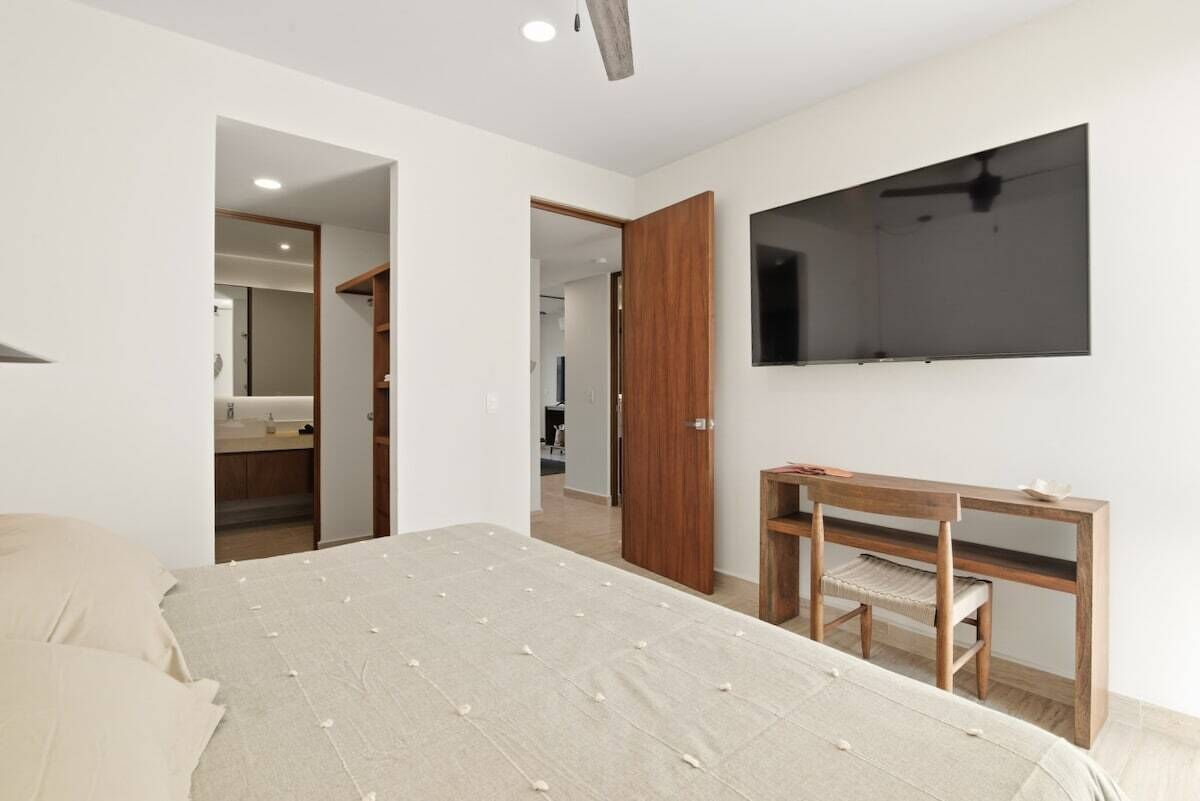 A bedroom with a bed, a wooden desk, a chair, a TV mounted on the wall, and a wardrobe.