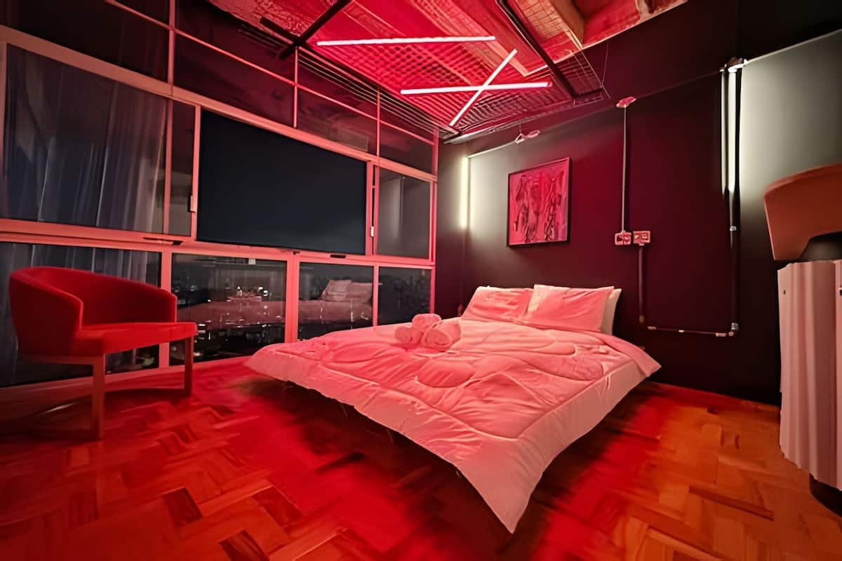 A modern bedroom with a large bed, a red chair, and a city view through large windows.