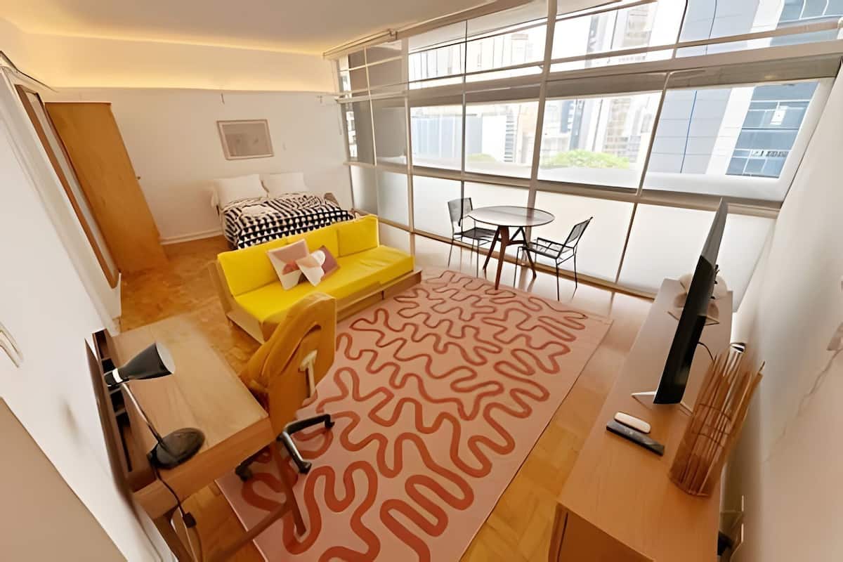 A modern living room with a yellow sofa, a patterned rug, a dining table with chairs, and large windows offering a view of buildings.