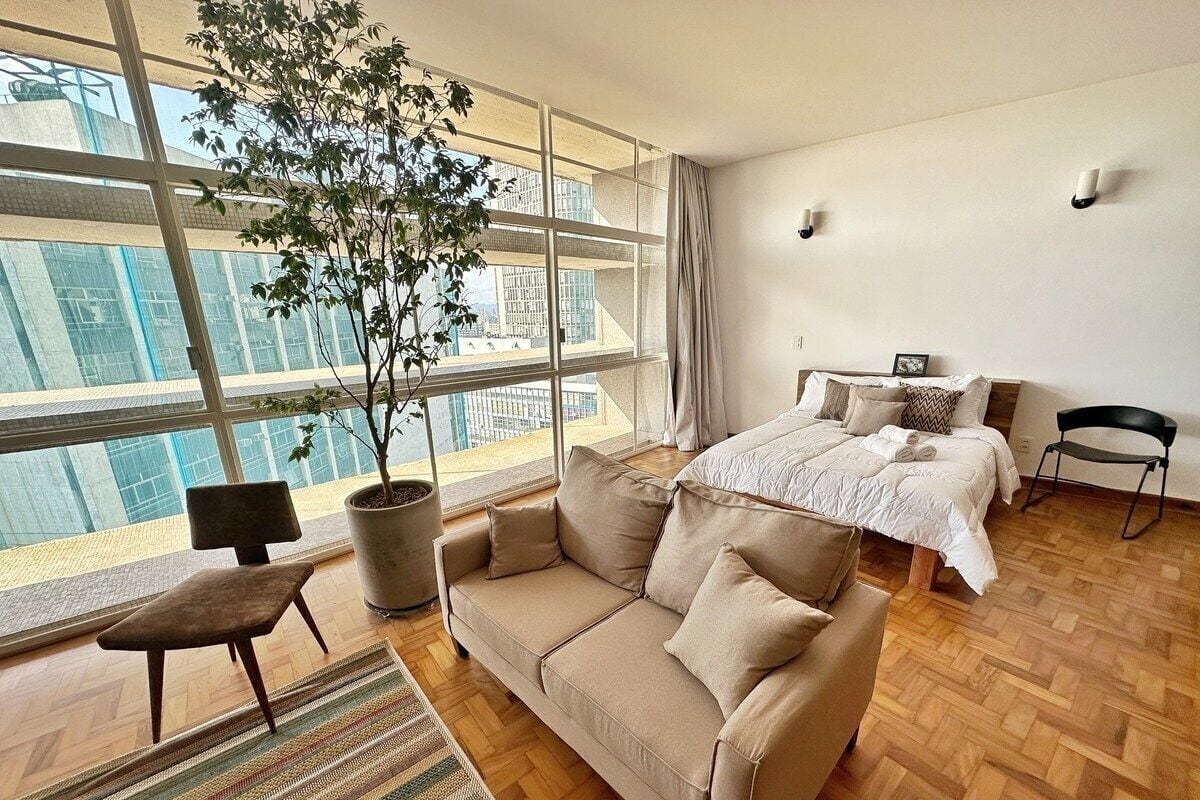 A modern hotel room with a large window, a sofa, a chair, a bed, and a potted plant.