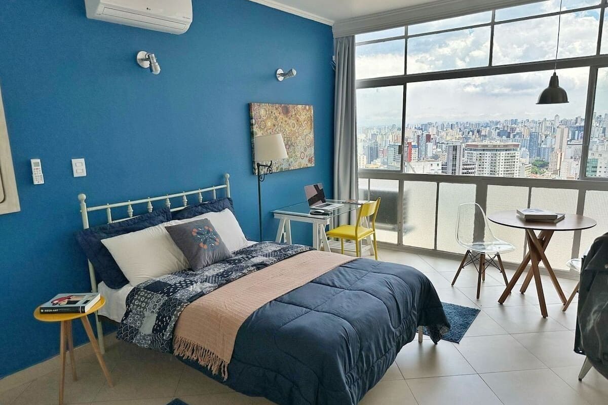 A bedroom with a bed, a desk, a chair, a window with a city view, and a balcony.