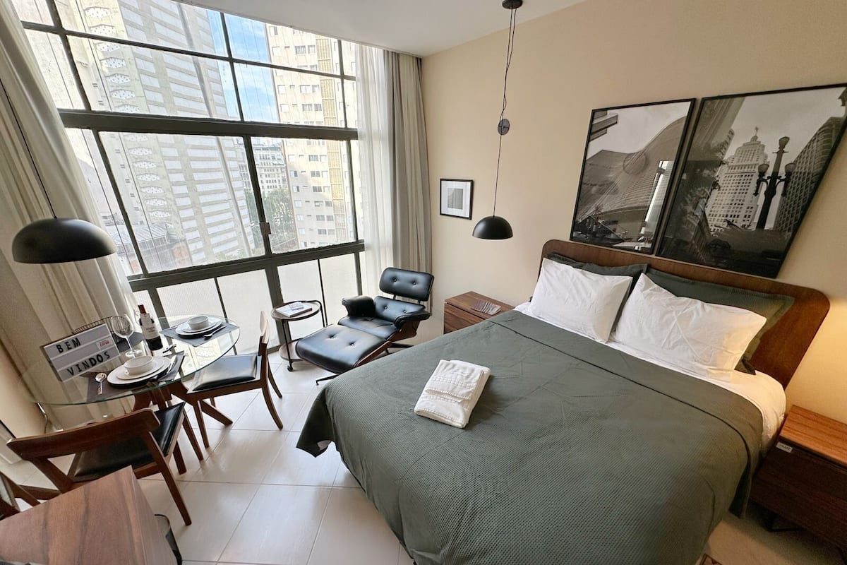 A hotel room with a large bed, a desk with a chair, a small table with a chair, a window with a view of buildings, and a painting on the wall.