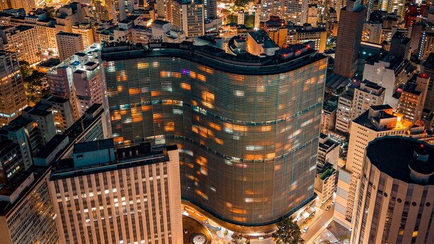 A high-rise building with a curved facade, surrounded by other tall buildings in an urban setting. - Vem pro Copan (São Paulo)