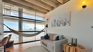A modern living room with a large window, a hammock, a white sofa, a round rug, and a wooden side table. - Vem pro Copan (São Paulo)
