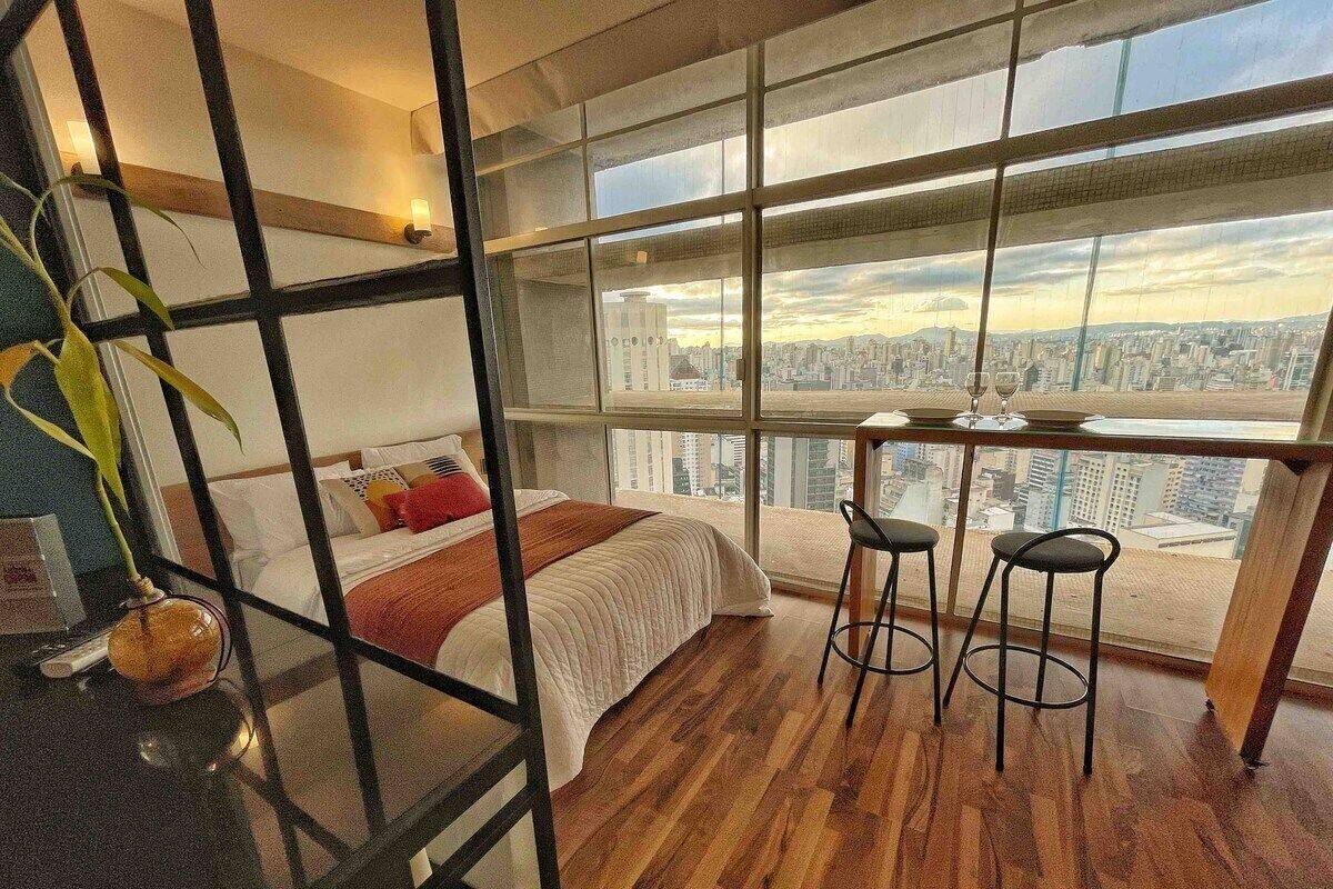 A modern hotel room with a large window, a bed, a small table with two stools, and a plant.