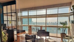 A spacious living area with a large window, a black sofa, a round coffee table, and a small kitchen area with stools. - Vem pro Copan (São Paulo)