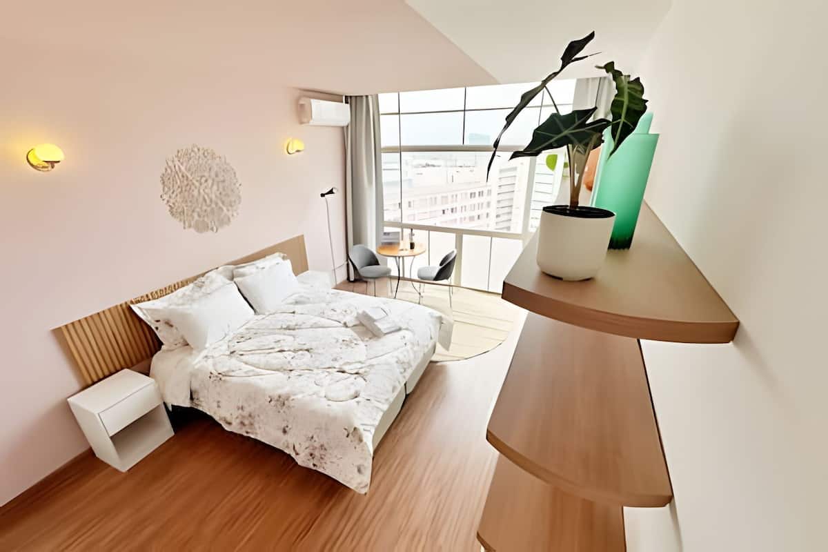 A modern bedroom with a wooden bed, a small bedside table, a large window with a view of buildings, and a green plant on a shelf.