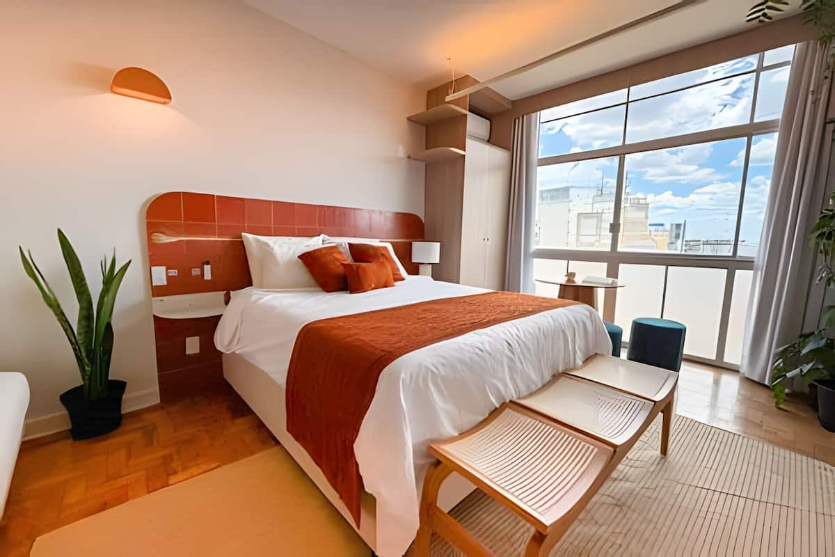 A modern hotel room with a large bed, a small table, and a view of the city through large windows.