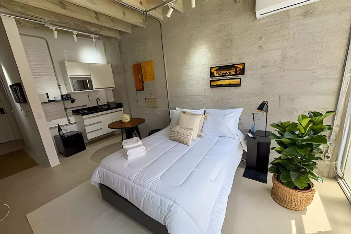A modern bedroom with a large bed, a small table, a potted plant, and a kitchen area visible through an open door.