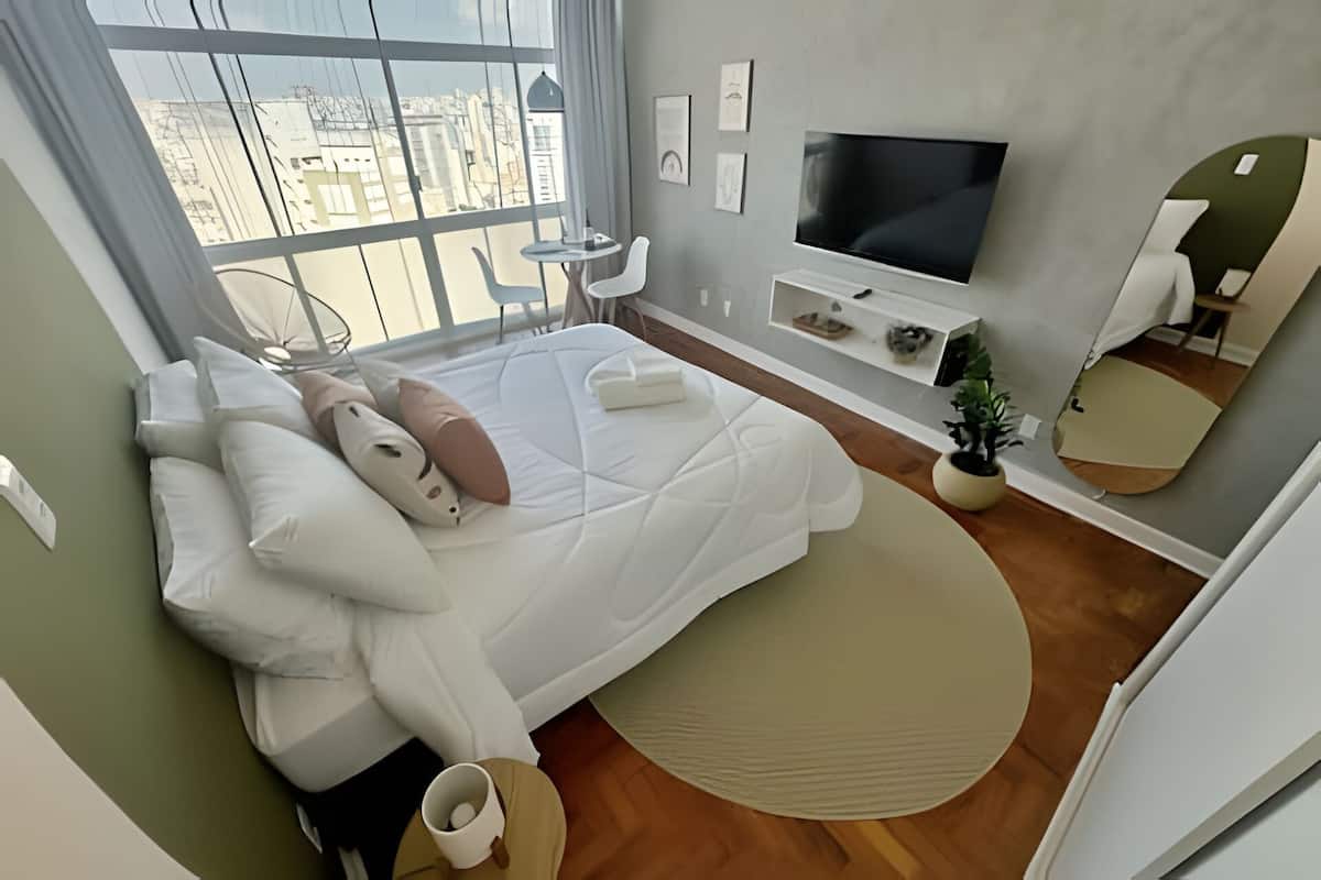 A modern bedroom with a large bed, a TV, a small table, and a view of the city through large windows.