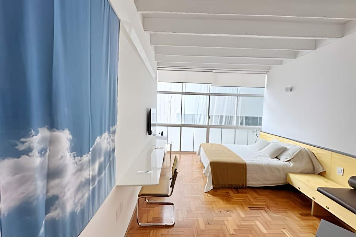 A bedroom with a bed, a desk, and a chair. There is a window with curtains, and a wall with a cloud mural.