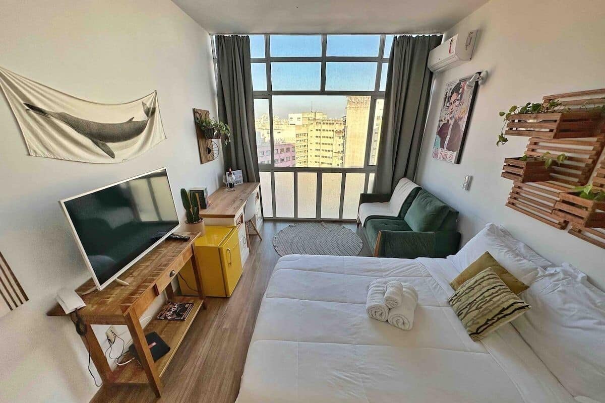 A hotel room with a bed, a TV, a desk, and a view of the city through large windows.