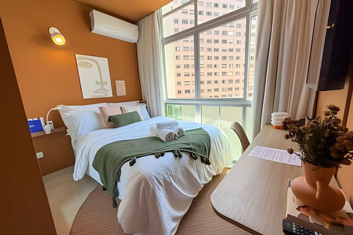 A hotel room with a bed, a desk, a window with a view of buildings, and an air conditioning unit.