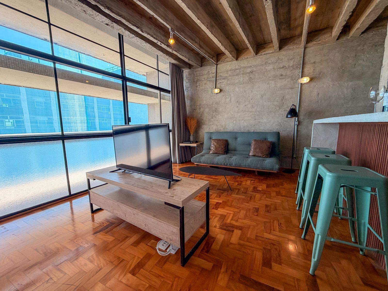 Una sala de estar moderna con un ventanal grande, piso de madera, un sofá, un televisor y taburetes de bar.