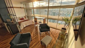 A high-rise apartment with a large balcony, a bed, a desk, and two armchairs. - Vem pro Copan (São Paulo)