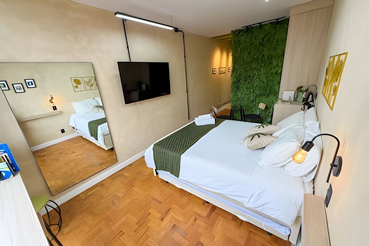 A hotel room with a large bed, a flat-screen TV mounted on the wall, a green wall feature, and a wooden floor.