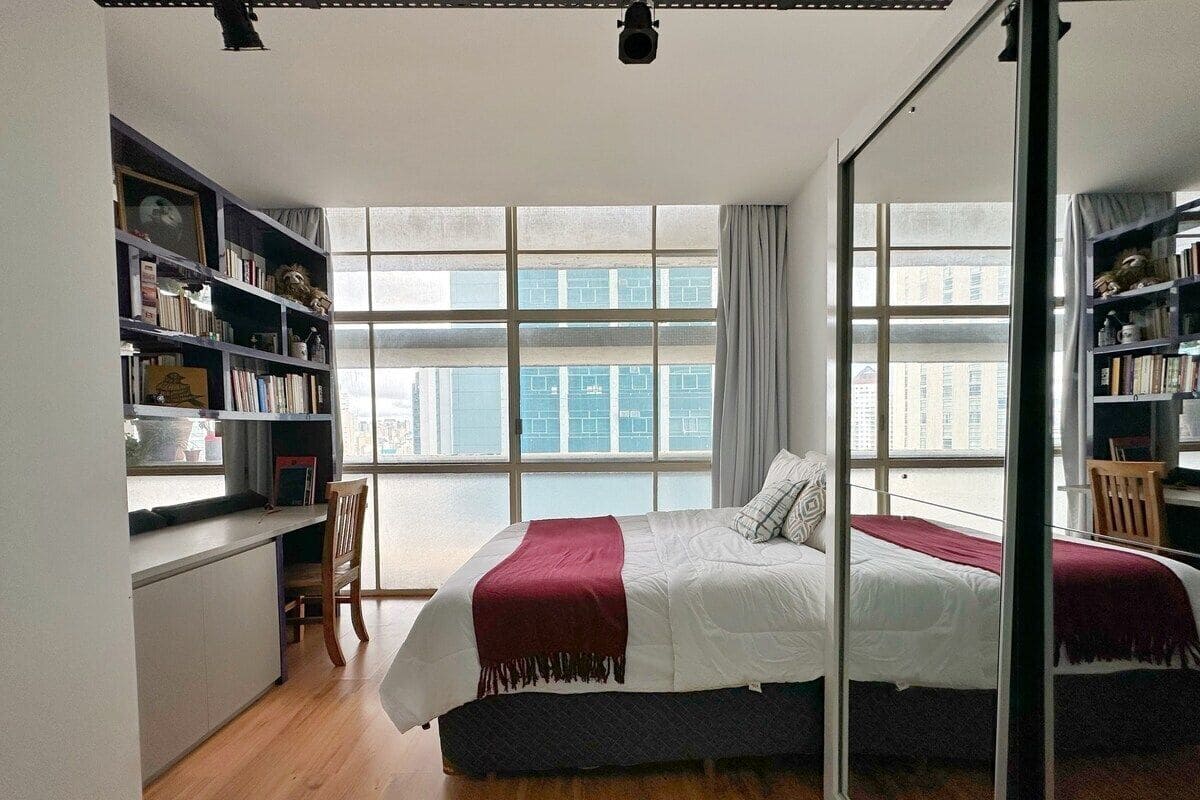 A bedroom with a large bed, a bookshelf, a desk, and a window with a view of buildings.