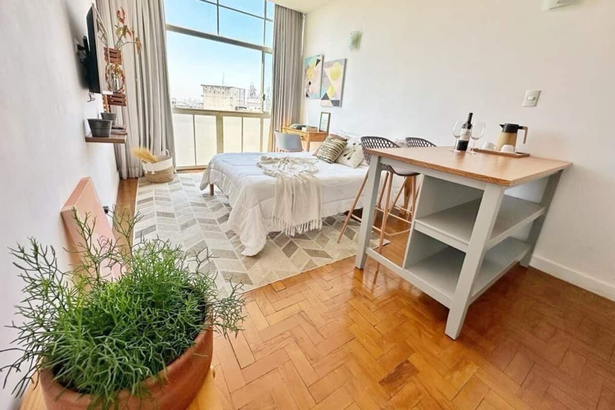 A modern hotel room with a large window, a bed, a desk, and a potted plant.