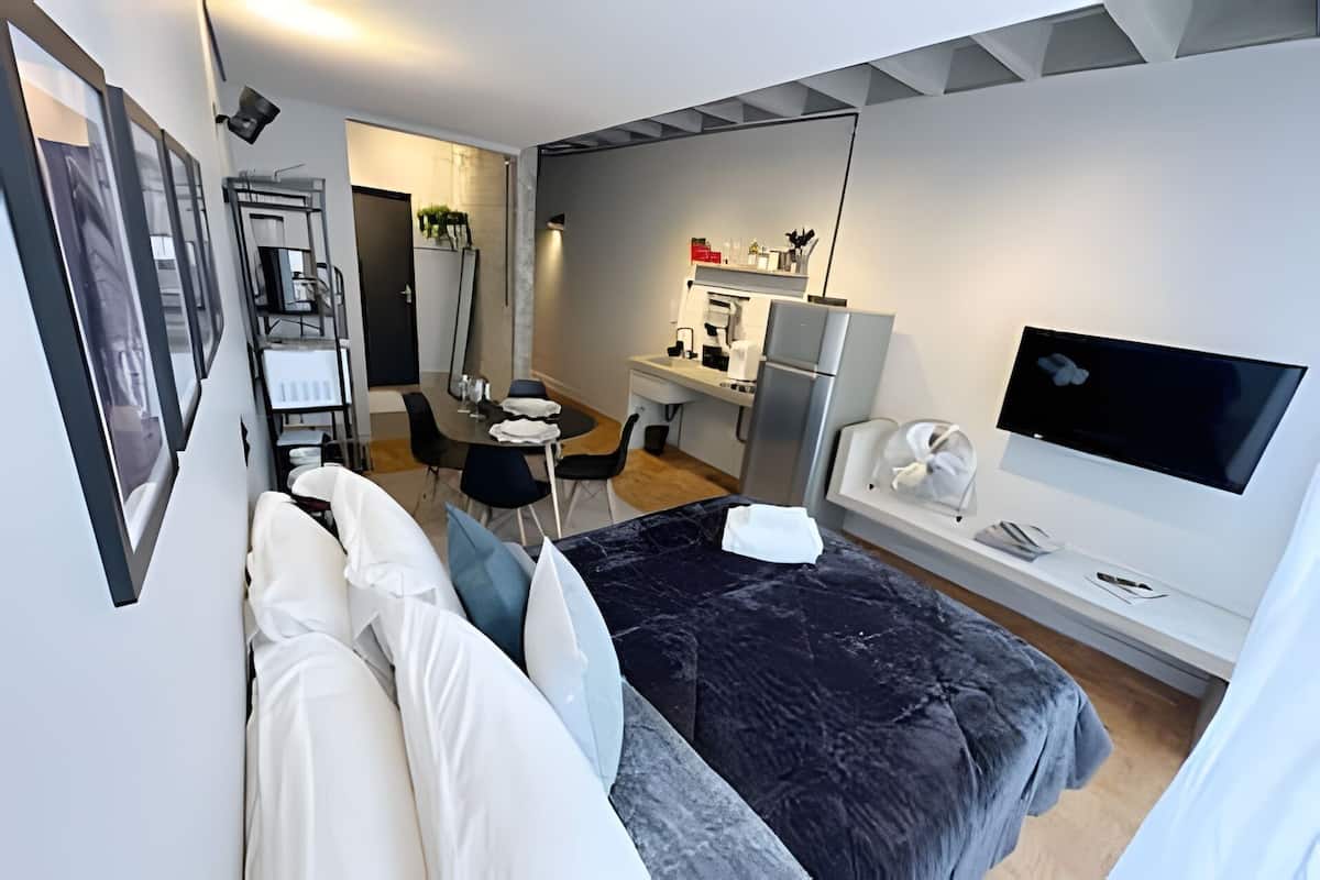A modern hotel room with a bed, a desk, a chair, a TV, and a kitchenette.