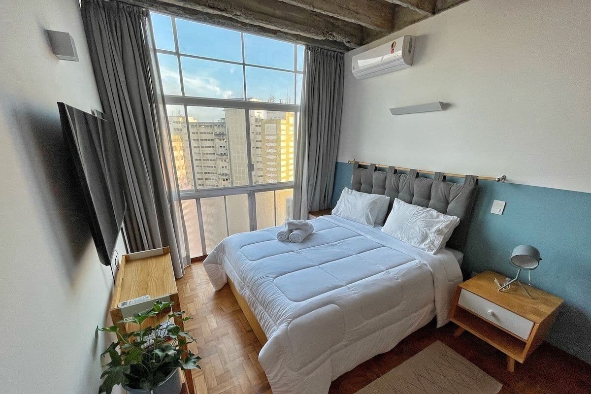 A modern bedroom with a large bed, a bedside table, a nightstand, a potted plant, and a view of buildings through the window.