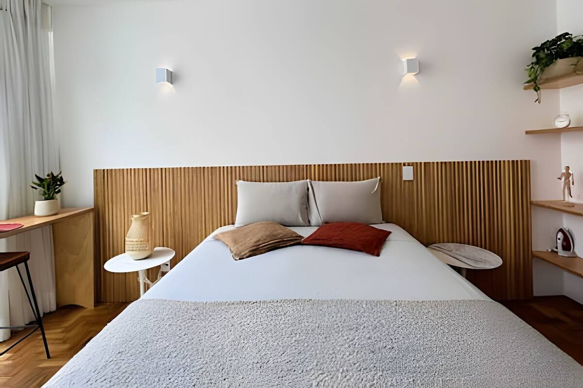 A modern hotel room with a wooden headboard, a large bed with multiple pillows, a small round table with a plant, and a shelf with decorative items.