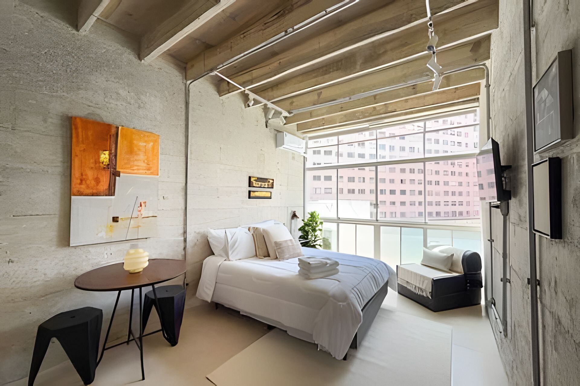 A modern bedroom with a bed, a small table, and a chair. There is a window with a view of buildings outside.