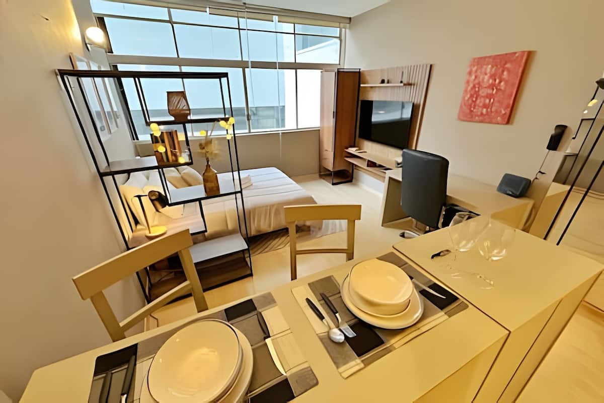 A modern hotel room with a bed, a desk, a chair, a TV, and a dining area with a table set for two.