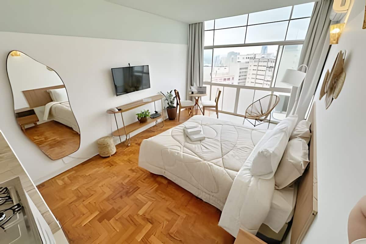 A modern bedroom with a large bed, a TV mounted on the wall, a wooden floor, and a city view through the window.