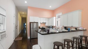 A kitchen with white cabinets, a stainless steel refrigerator, a microwave, and a sink. There are four stools at the island. - Cute and Cozy 2BR/2BA w/Parking, Yard, King Suite & Blocks to Local Favorites (New Orleans)
