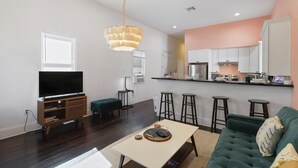 A modern living room with a flat-screen TV, a green sofa, a coffee table, and a kitchen area with bar stools. - Cute and Cozy 2BR/2BA w/Parking, Yard, King Suite & Blocks to Local Favorites (New Orleans)