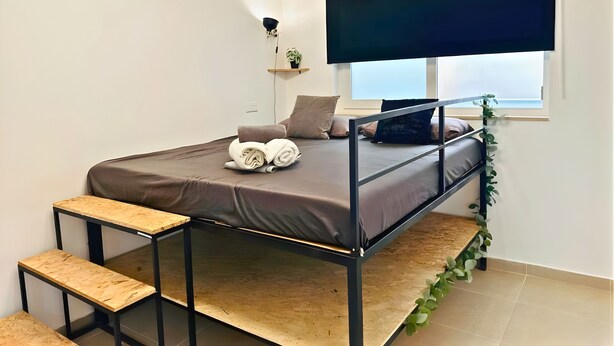 Een kleine kamer met een stapelbed, een tafeltje, een plank met een plant en een raam met uitzicht.