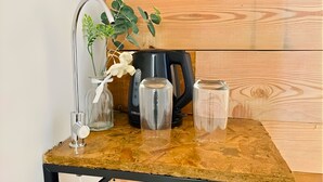 Een houten tafel met een koffiezetapparaat, twee glazen recipiënten en een vaas met een plant en witte bloemen.